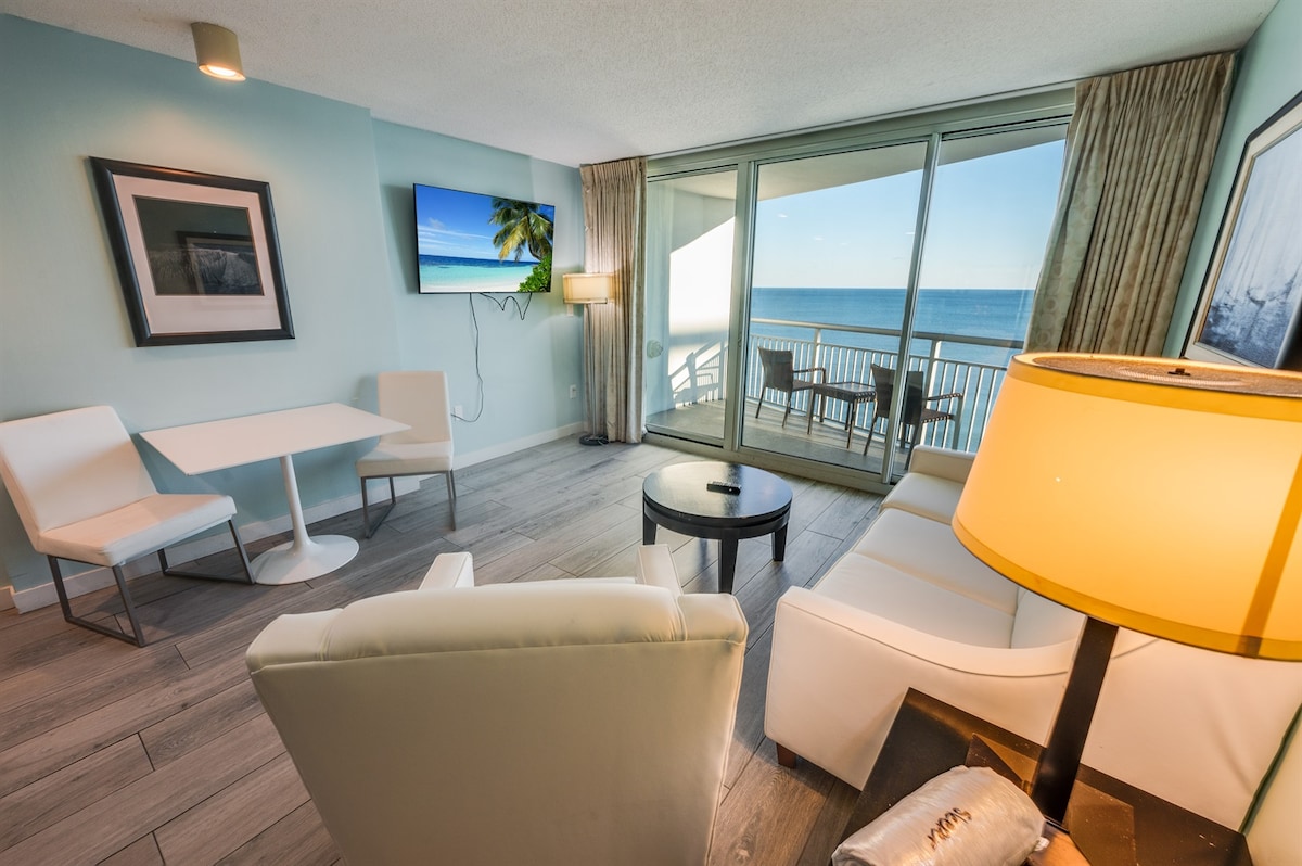 A modern living area features a comfortable seating arrangement with a light-colored sofa and armchair. A sleek coffee table is positioned near a small dining table with two chairs. Large glass doors provide access to a private balcony, showcasing ocean views.