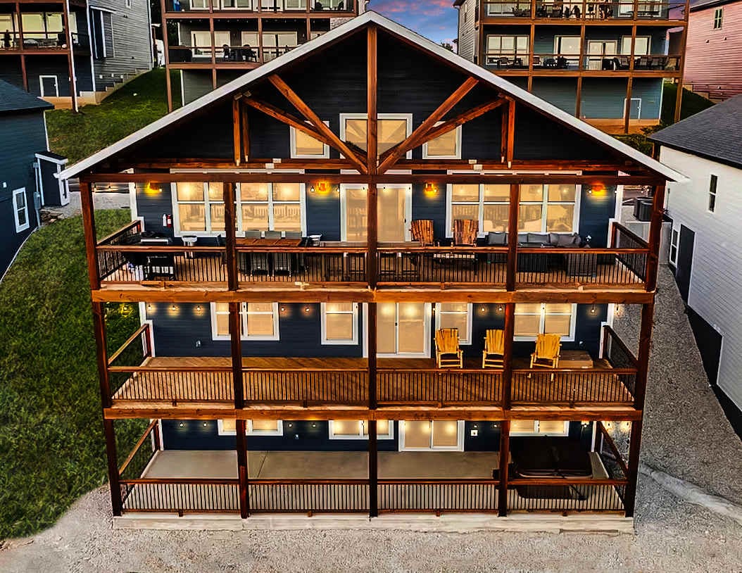 The multi-level lodge features a spacious wooden exterior with large balconies. Multiple seating areas can be seen on each level, showcasing wooden chairs and railings. The structure is set against a hillside, highlighting its expansive design and inviting atmosphere.