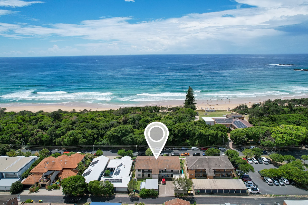 Aerial view of the beachfront property showcases the ocean's waves meeting the sandy shore, with local vegetation lining the coastline. Nearby buildings are visible, with a clear view of the vibrant blue sea and beachgoers in the distance.