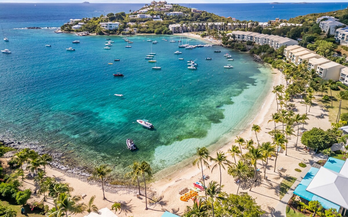 An aerial view captures a serene beach with soft, sandy shores and turquoise waters. Small boats gently float in the bay, surrounded by lush greenery and palm trees. Resort-style buildings line the coast, providing a harmonious backdrop to the tranquil seaside landscape.