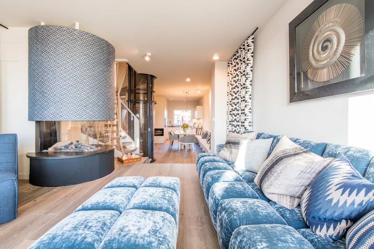A spacious living area is highlighted by a plush blue sectional sofa and a gas fireplace, which serves as a centerpiece. A stylish spiral staircase leads to upper levels, while natural light floods the space through large windows.