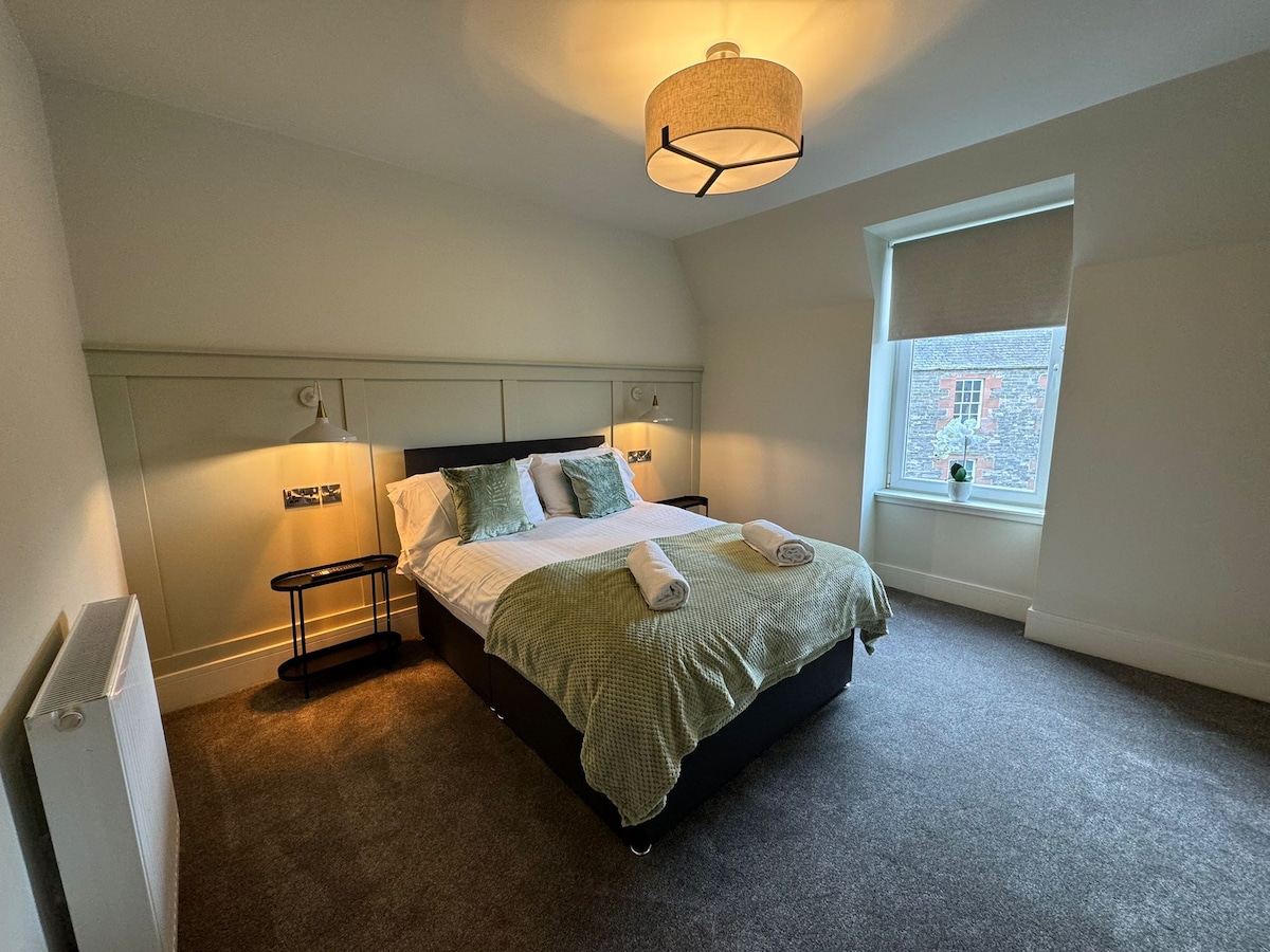 A spacious bedroom features a comfortable bed with soft bedding and decorative pillows. Natural light filters through a window, illuminating the room's neutral tones. A bedside table and lamp complete the space, providing functionality and warmth.