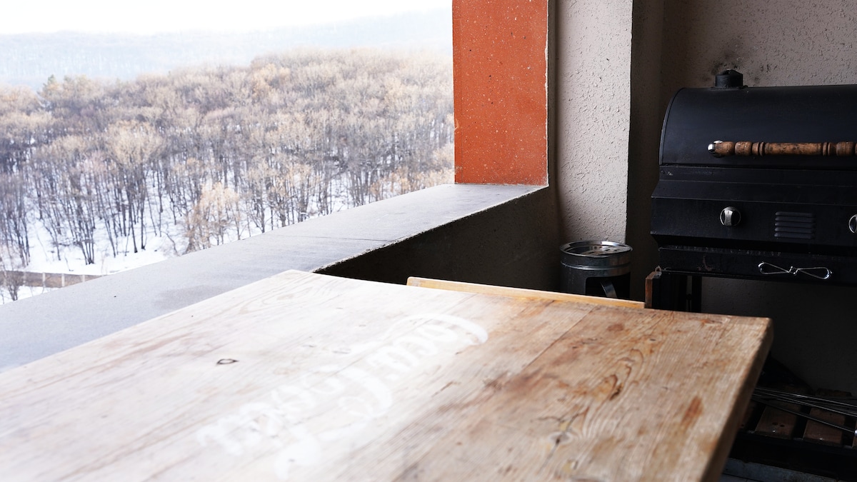 A wooden table is positioned on a private balcony, complemented by a barbecue grill nearby. The scenic view features snow-covered landscapes and trees, providing a serene outdoor space for relaxation.