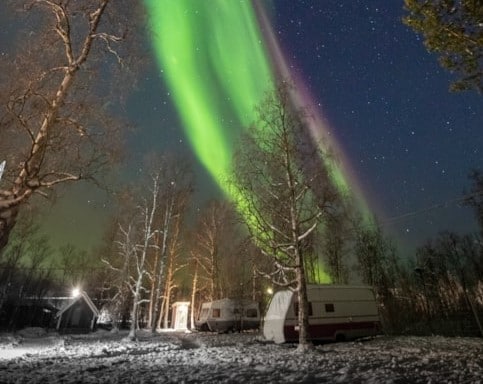 The night sky is illuminated by vibrant green auroras, stretching across a backdrop of stars. Snow-covered trees frame the scene, with two parked trailers visible in the foreground, enhancing the serene winter atmosphere.