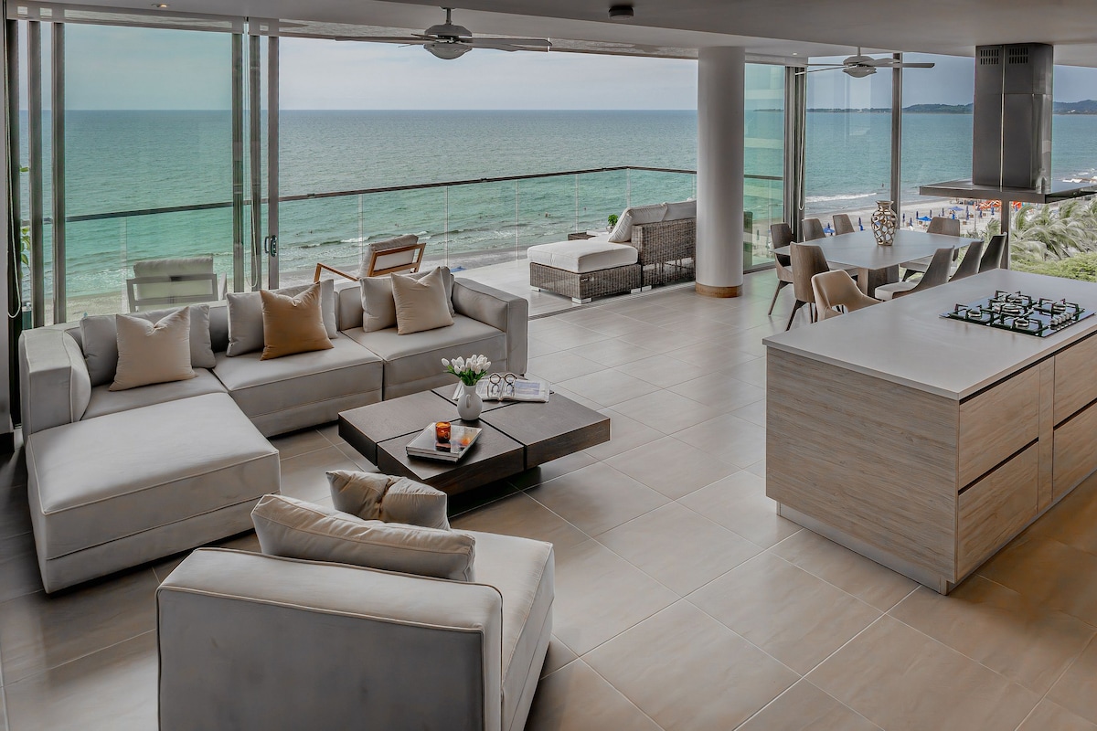 A spacious living area features large glass windows, offering a panoramic view of the sea. Comfortable sofas surround a modern coffee table, inviting relaxation. An open kitchen with a central island and contemporary design is visible, enhancing the bright and airy ambiance of the space.