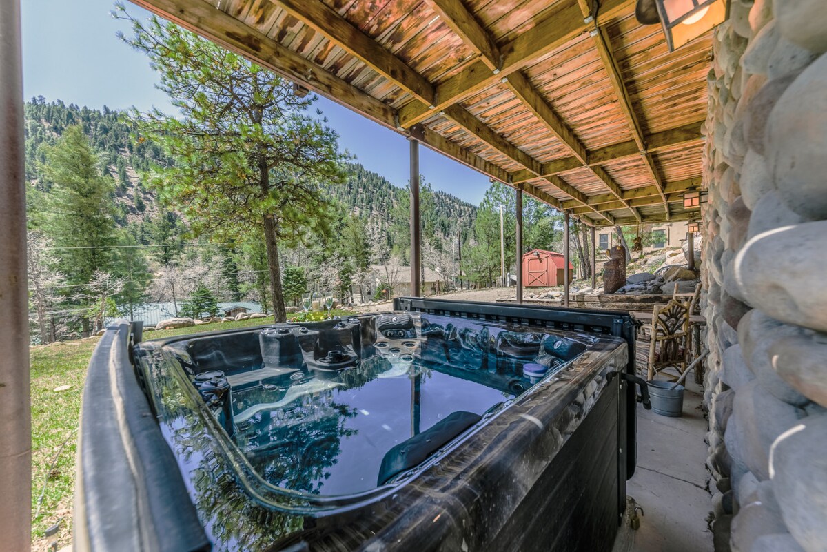 A private outdoor hot tub is located on a covered deck, surrounded by tall trees and mountain scenery. The tranquil setting invites relaxation, with views of the serene landscape visible in the background. Natural light enhances the space, creating a peaceful atmosphere.