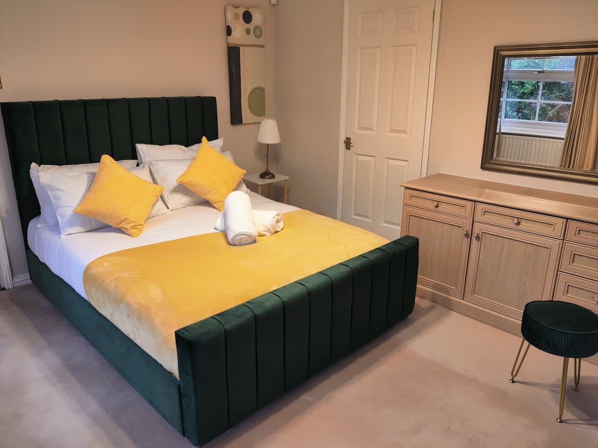A spacious bedroom is furnished with a plush bed featuring a green upholstered headboard and a bright yellow throw. Cushions are neatly arranged at the head of the bed. A wooden dresser complements the room, alongside a circular stool and a mirror reflecting natural light from a nearby window.