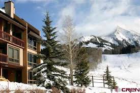 The exterior of the condo is shown, featuring a three-story building with wooden balconies. Snow-covered trees enhance the winter landscape, and majestic mountains are visible in the background, highlighting the property's scenic location.