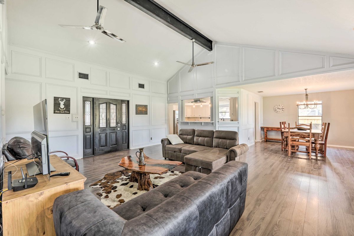 An open living area features a comfortable sectional sofa, a wooden coffee table, and a Smart TV. Natural light fills the space through large windows, which are accented by ceiling fans and modern light fixtures. An adjoining dining area is visible in the background.
