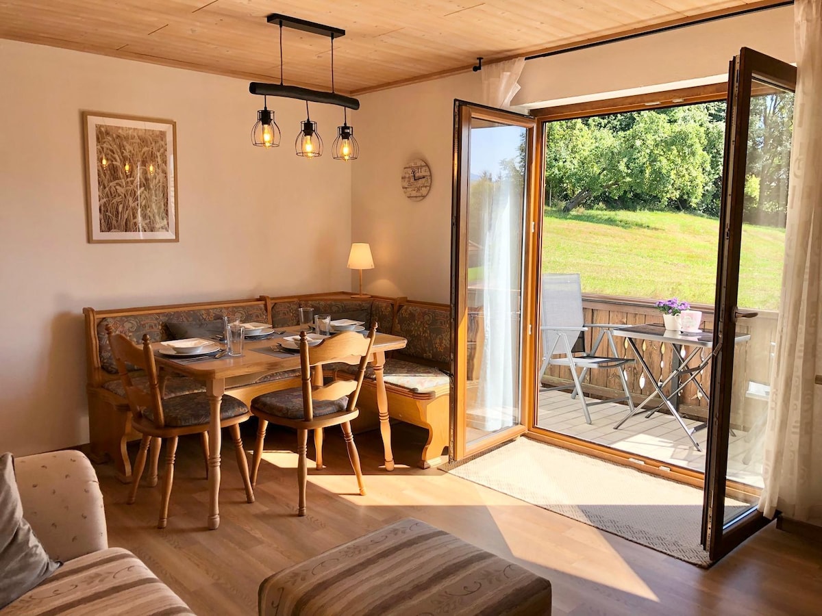 A cozy dining area features a wooden table surrounded by padded chairs, ideal for family meals. Large glass doors open onto a balcony, allowing natural light to fill the space. A softly glowing lamp and tasteful artwork provide a welcoming touch.