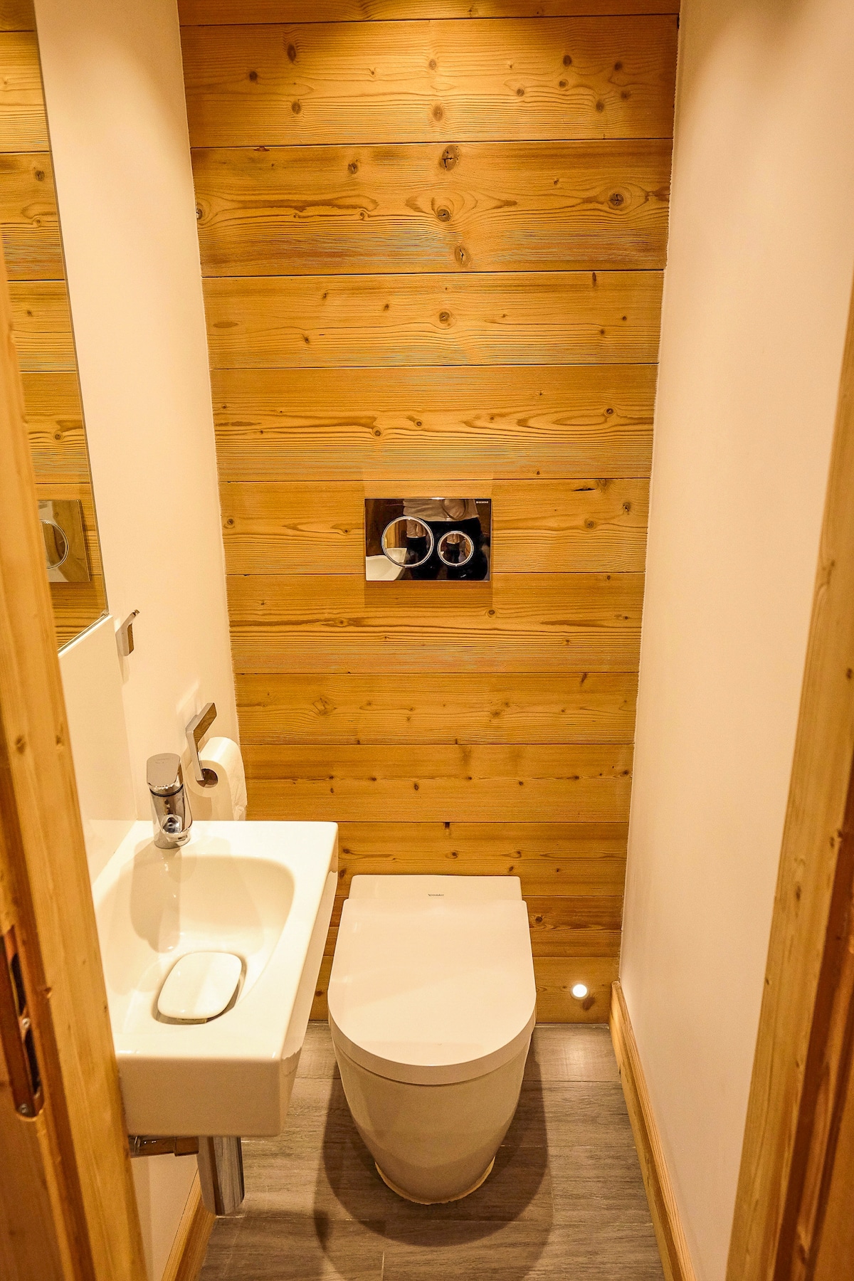 A compact restroom features a modern toilet and a sleek sink with a mirror above. The wooden wall adds warmth to the space, while subtle lighting highlights the floor tiles.