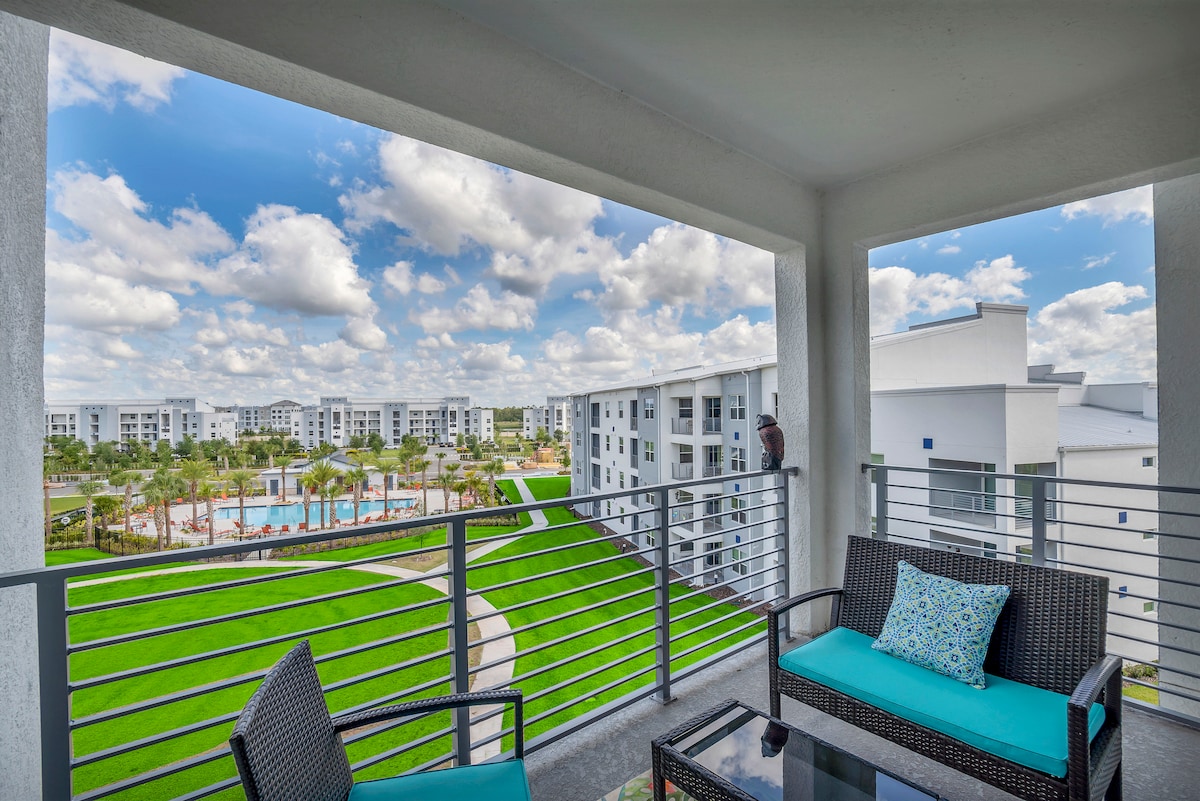 A private balcony features outdoor seating with cushions, overlooking a landscaped area with a shared pool surrounded by greenery. The view includes multiple buildings in the distance against a backdrop of a partly cloudy sky.