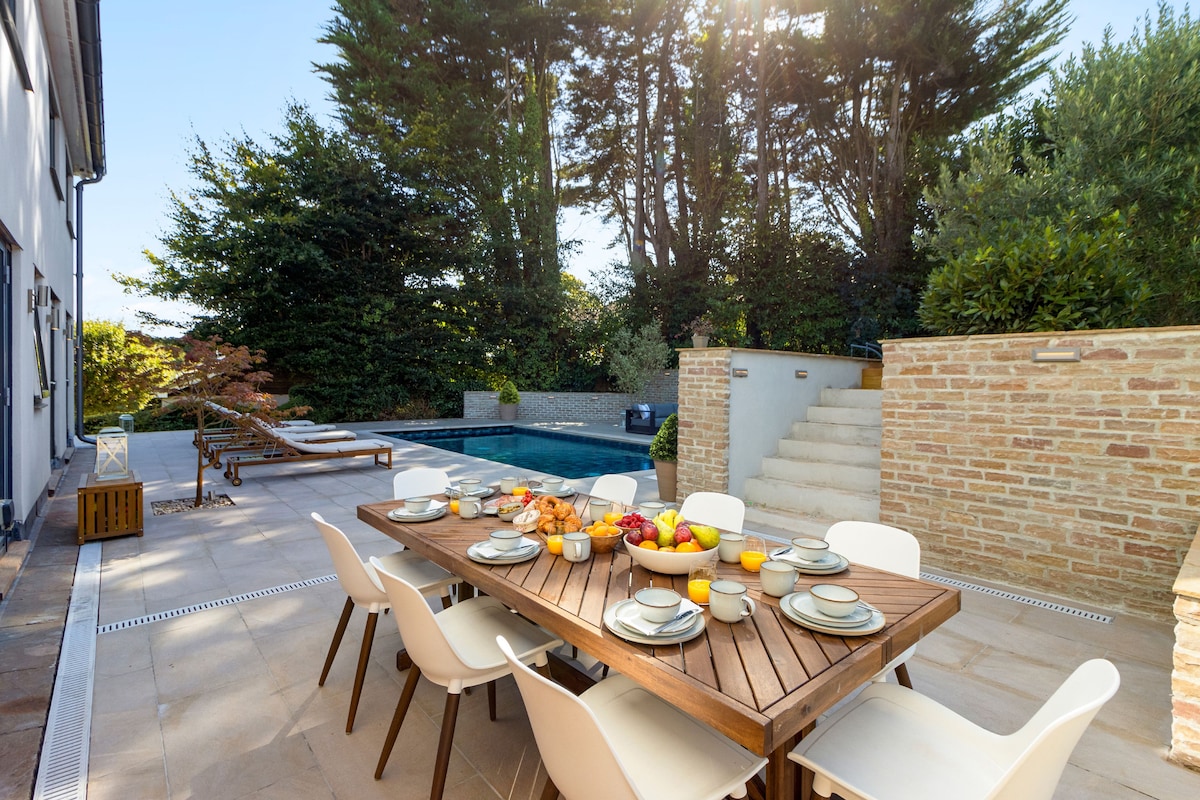 An outdoor dining area features a large wooden table set with plates, glasses, and colorful fruit. Surrounding greenery and sunlit trees create a natural backdrop, while a heated pool and lounge chairs are visible in the distance, inviting relaxation and al-fresco meals.