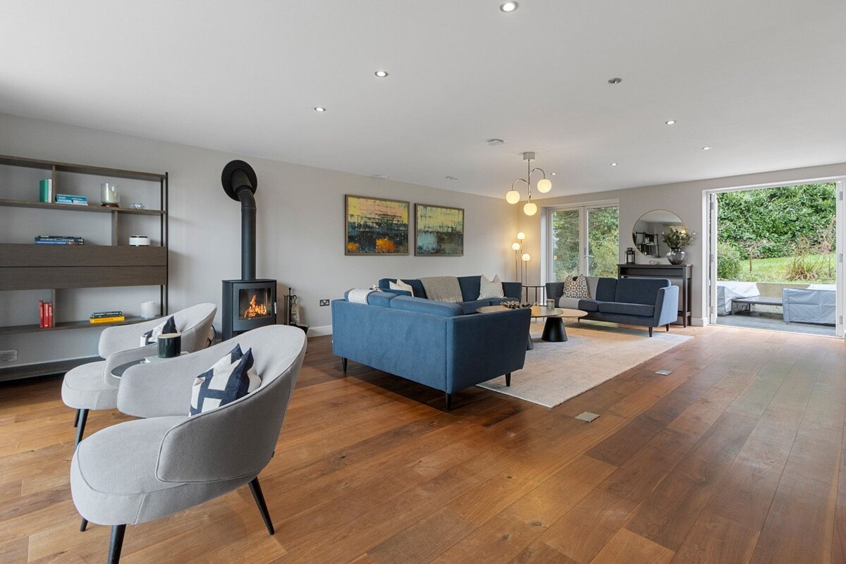 A spacious living area is presented with sleek furnishings, featuring cozy sofas and armchairs arranged around a modern woodburner. Large sliding doors provide access to the outdoor space, inviting natural light and views of the garden.