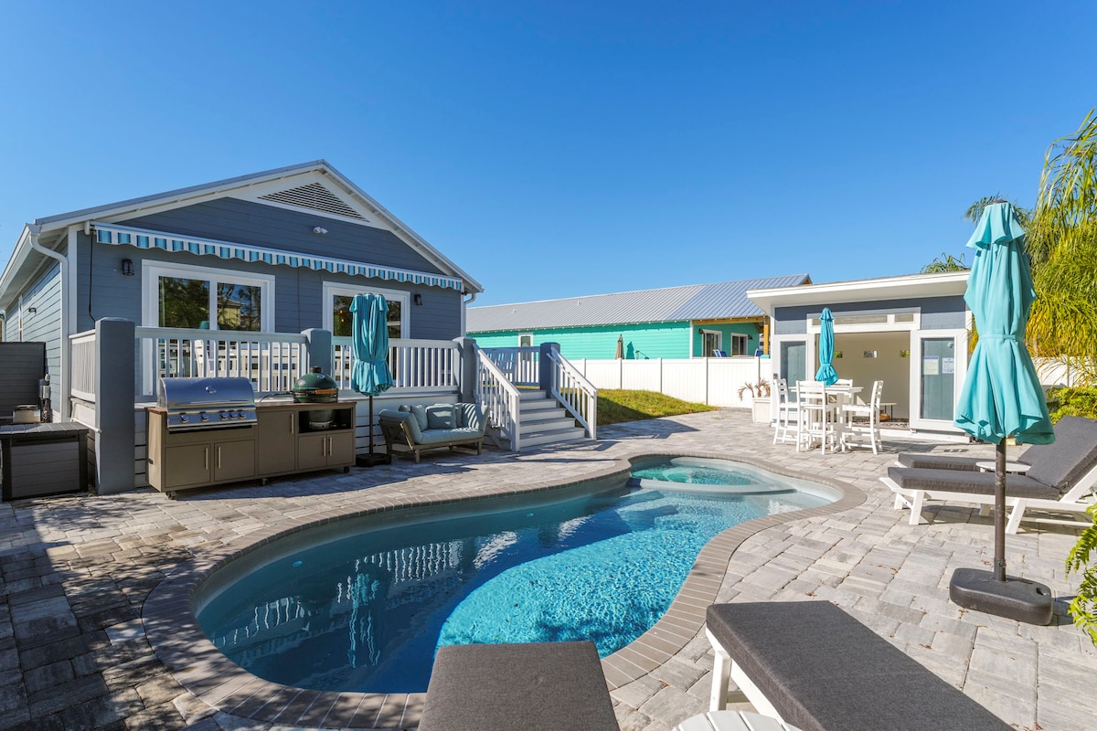 A welcoming outdoor area features a pool surrounded by paved stones and lounge chairs. A grilling station is positioned nearby, and vibrant umbrellas provide shade. In the background, a light-colored cabana is visible, completing the coastal retreat atmosphere.