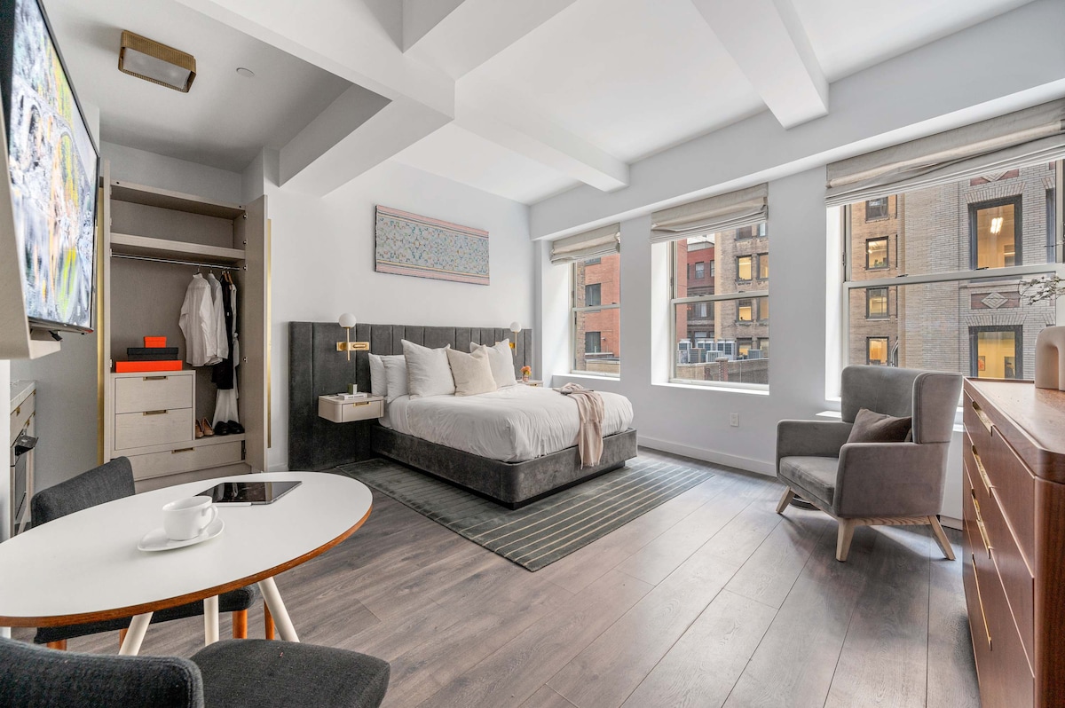 A well-lit studio features a spacious king bed against a dark upholstered headboard. Natural light streams through large windows, illuminating a small dining table and a cozy chair. A modern dresser and closet provide ample storage, enhancing the inviting atmosphere of the space.