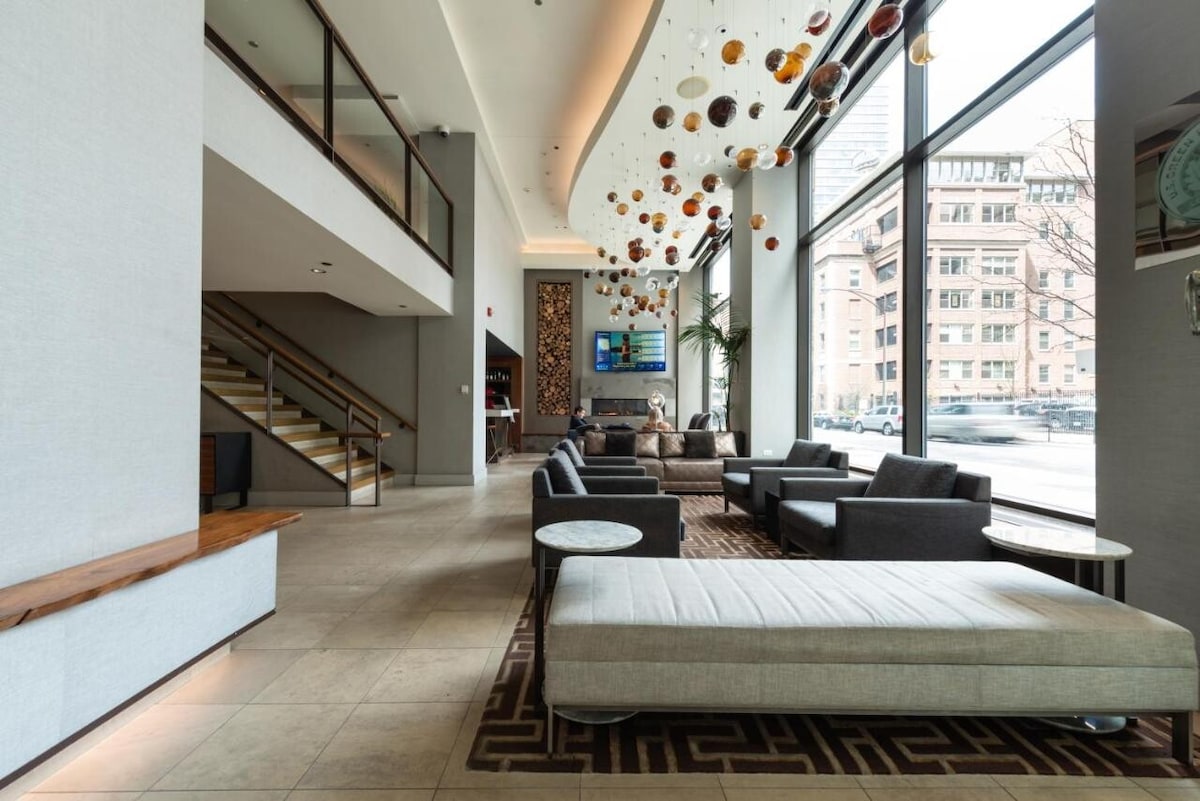 A spacious hotel lobby is highlighted by large windows allowing natural light to fill the area. Comfortable seating arrangements with gray sofas and armchairs are positioned on a patterned rug. Unique decorative elements hang from the ceiling, contributing to the contemporary design.