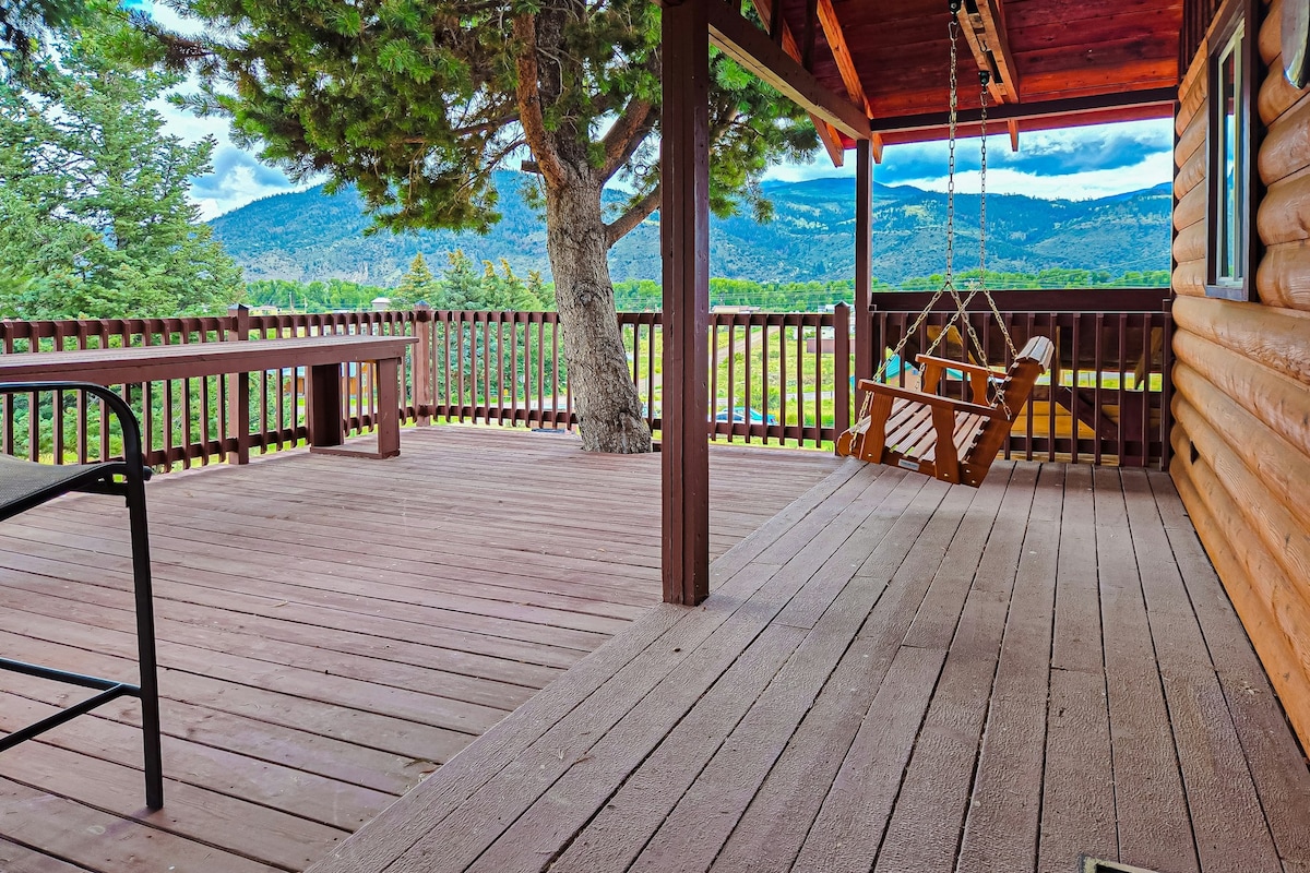 A spacious wooden deck is presented, featuring a swing chair and a large table. The surrounding railings are visible, alongside lush trees and mountain views in the background. Natural light highlights the deck’s warm tones, creating an inviting outdoor space.