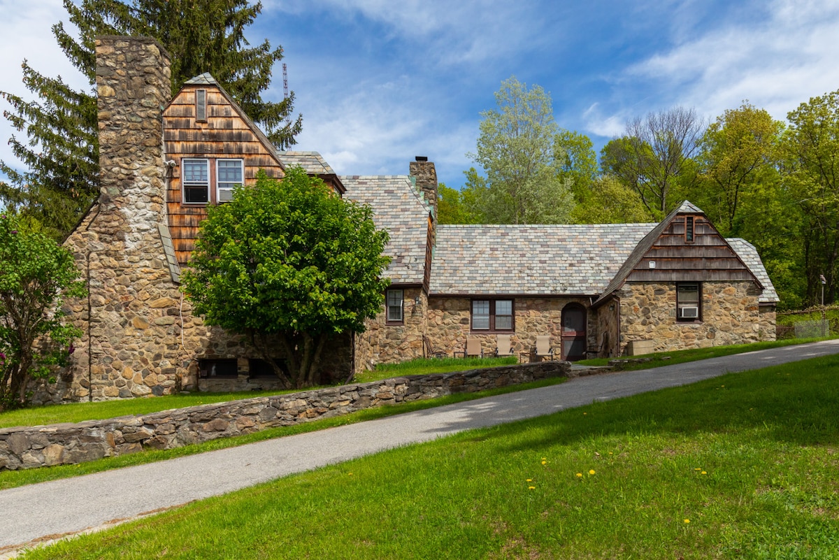 The historic stone Gate House features a blend of rustic stone walls and wooden shingles. The facade is framed by lush greenery, and the gentle slope of the lawn leads to a paved pathway. A cozy seating area is visible near the entrance.