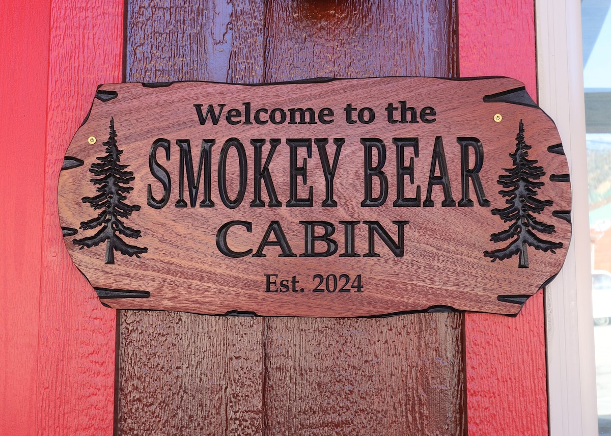 A carved wooden sign welcomes guests to the Smokey Bear Cabin, featuring the cabin name in bold letters. The sign is adorned with tree silhouettes, resting against a rustic wooden door painted in a vibrant red hue.