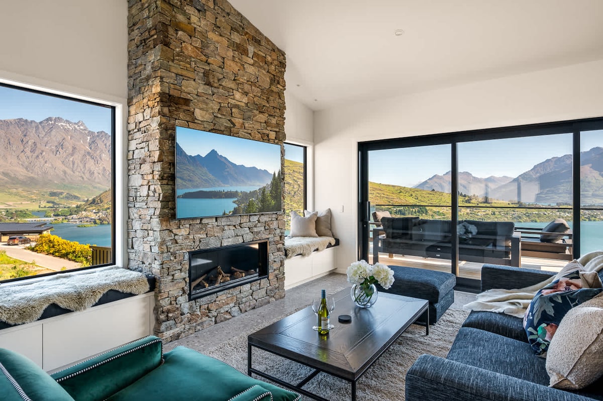 A modern living area showcases a natural stone fireplace and substantial windows framing stunning views of Lake Wakatipu and the surrounding mountains. A flat-screen TV is mounted above the fireplace, while a stylish coffee table is complemented by comfortable seating arrangements, creating a relaxed atmosphere.