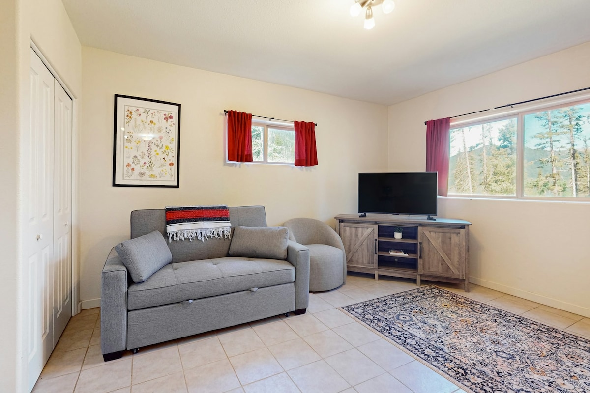 A living area features a grey sofa bed and a sitting chair, complemented by a wooden media console with a television. Natural light enters through two windows adorned with red curtains, and a decorative area rug adds warmth to the tiled floor.