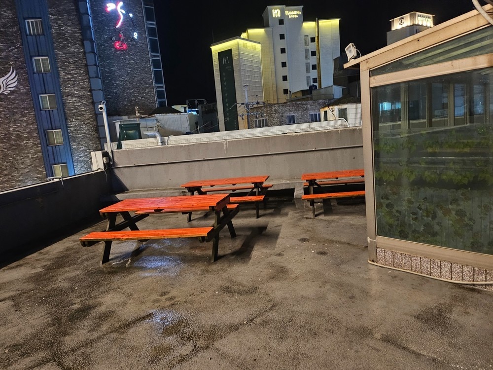 A rooftop area features several wooden picnic tables with orange-painted surfaces. The ground is wet, reflecting the surrounding urban buildings lit at night, creating a lively atmosphere. The glass enclosure at the edge indicates space for gatherings, with bright lights from nearby structures illuminating the area.