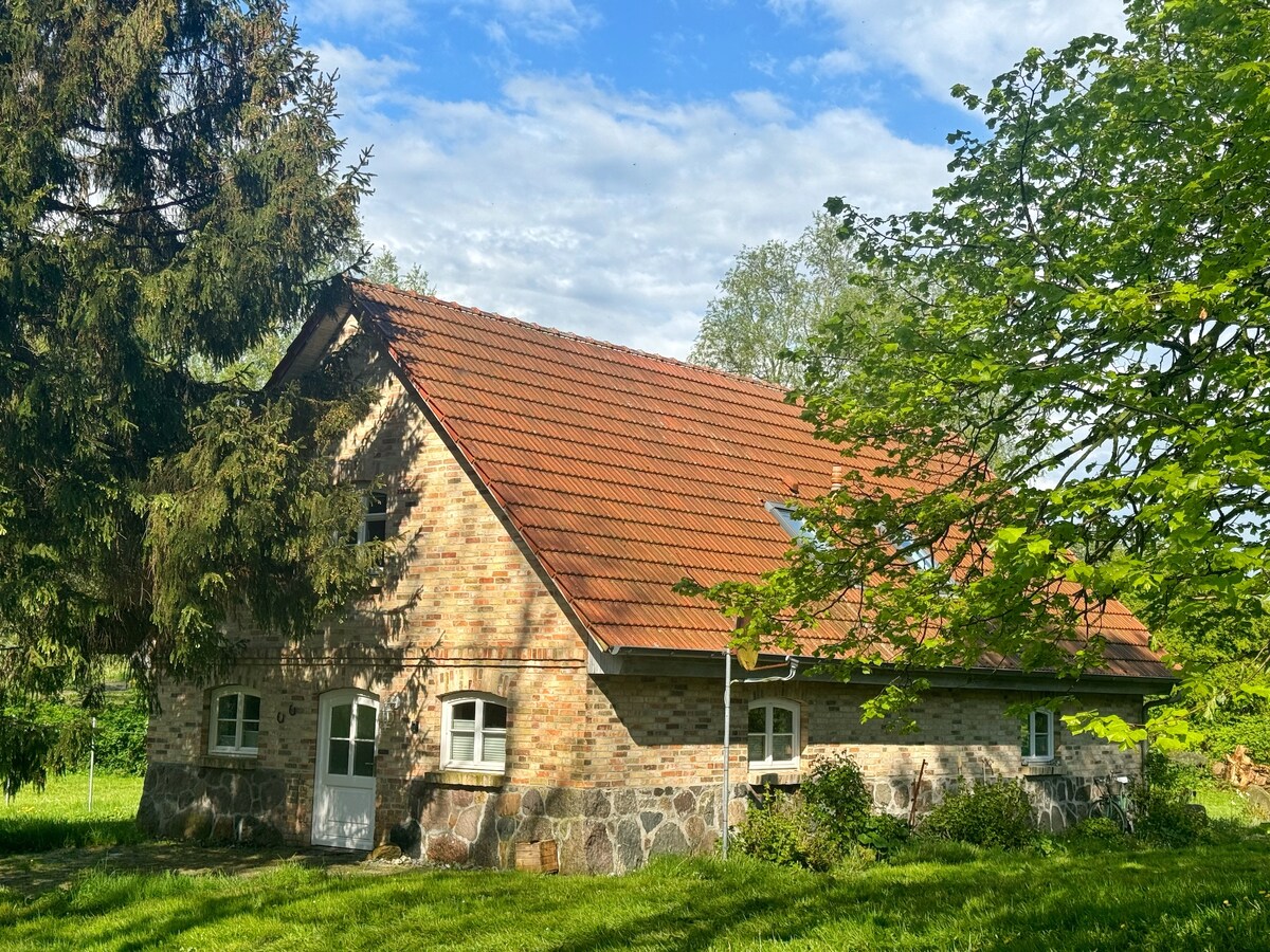 The two-story brick house features a distinctive red tiled roof, complemented by surrounding greenery. Large windows provide natural light to the interior. An expansive lawn leads up to the entrance, framed by mature trees and shrubs, creating a peaceful and natural environment.