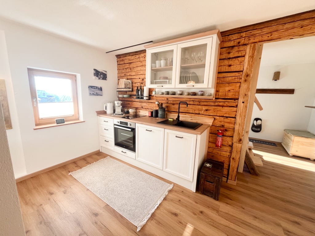 A rustic kitchen space features wooden wall panels that complement white cabinetry. An oven, stove, and sink are neatly arranged along the wall, with dishes and utensils displayed in glass-fronted cupboards. A window allows natural light to brighten the area, adding warmth to the design.