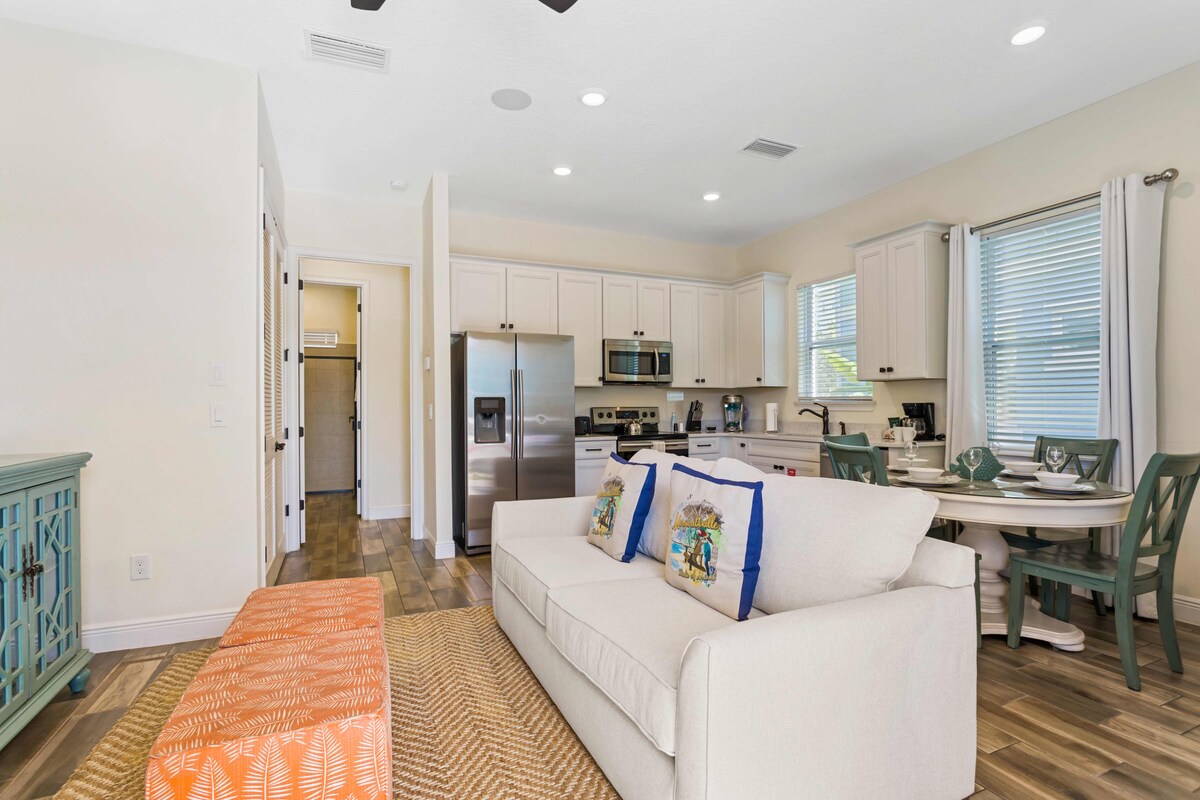The bright living area features a plush white sofa with decorative pillows and an orange accent ottoman. An open-concept kitchen includes modern stainless steel appliances and a dining table for four, all surrounded by natural light filtering through the windows.