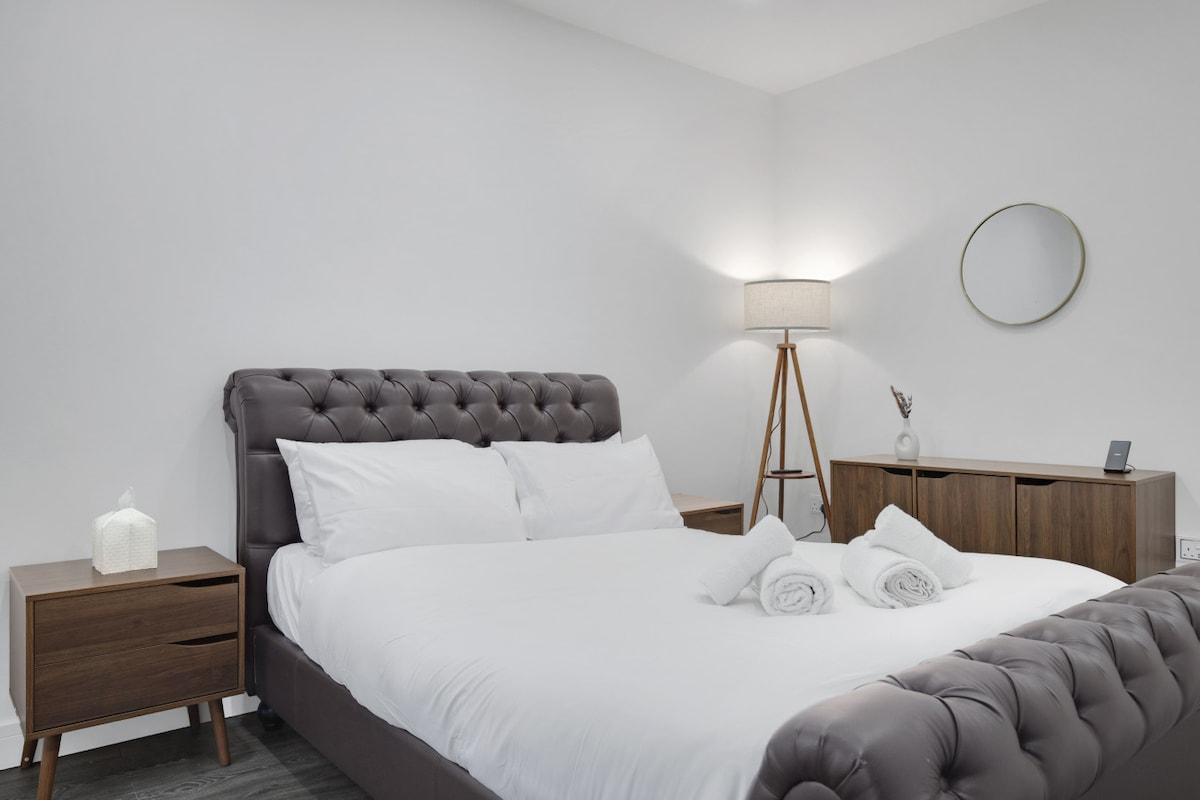 A well-appointed bedroom is designed for comfort, featuring a spacious double bed with tufted upholstery. Bedside tables flank the bed, and a light source emanates from a stylish lamp. A large sideboard adds storage, while a round mirror is mounted on the wall.