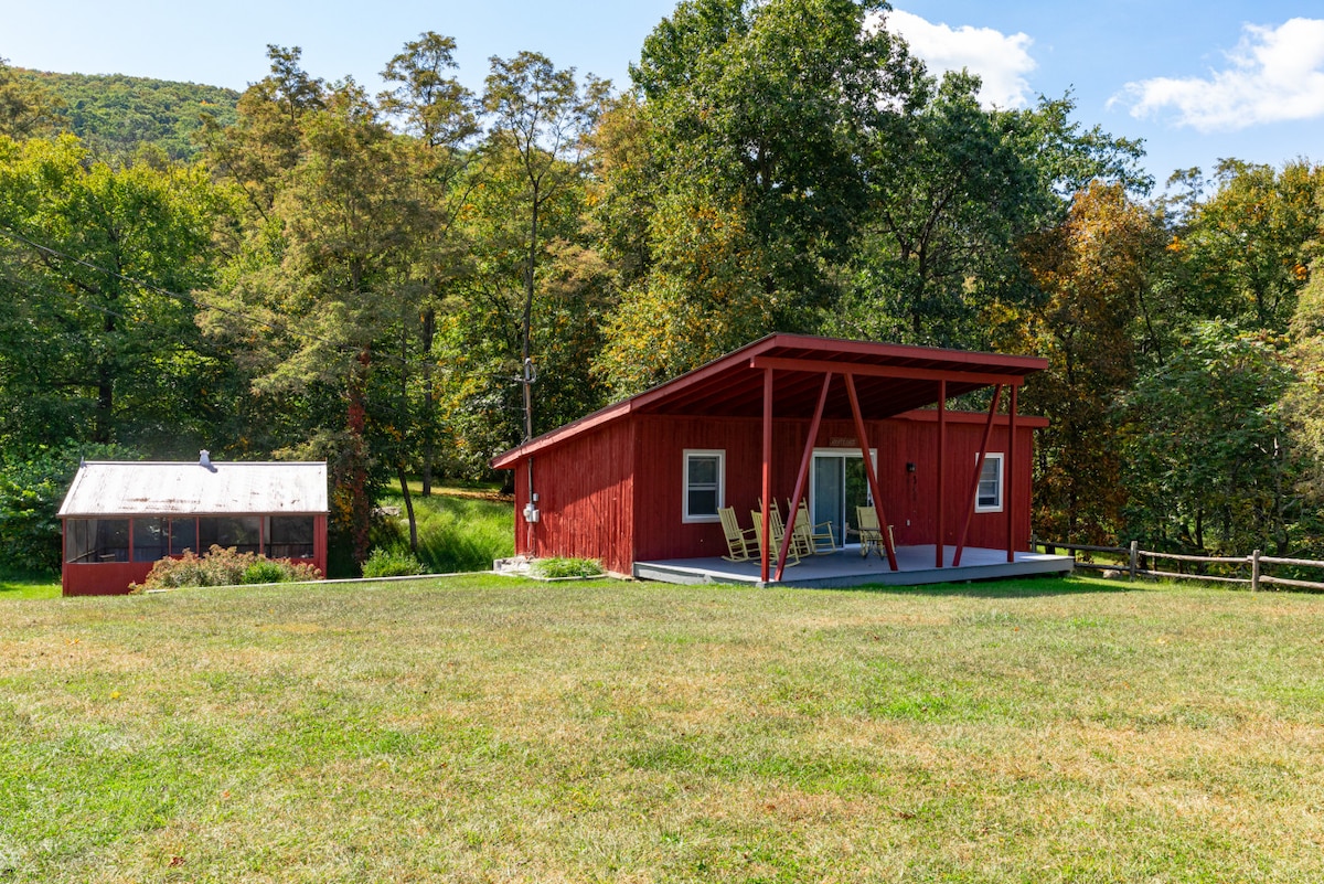 A red cottage is positioned against a backdrop of trees, showcasing a spacious covered porch with rocking chairs. Adjacent to the cottage is a separate screened house, all set on a grassy area with gentle hills in the distance.