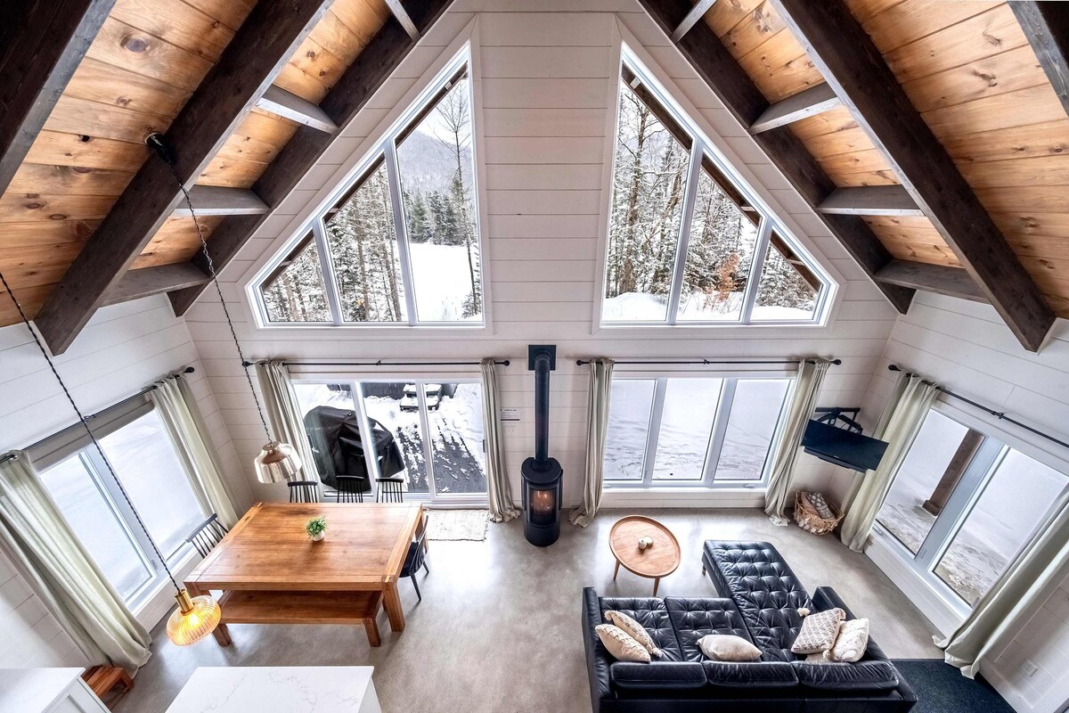 An airy living space features a high, triangular ceiling with exposed wooden beams. Large windows allow natural light to flood the area, showcasing views of the surrounding snow-covered landscape. A dining table sits adjacent to a cozy seating arrangement, complemented by a modern fireplace.