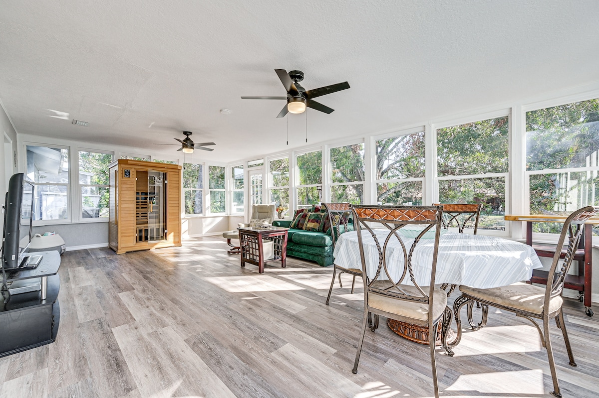 A bright sunroom features large windows allowing natural light to fill the space. Several seating areas are present, including a round table with chairs and a loveseat. A wooden sauna unit is positioned in one corner, complementing the inviting atmosphere.