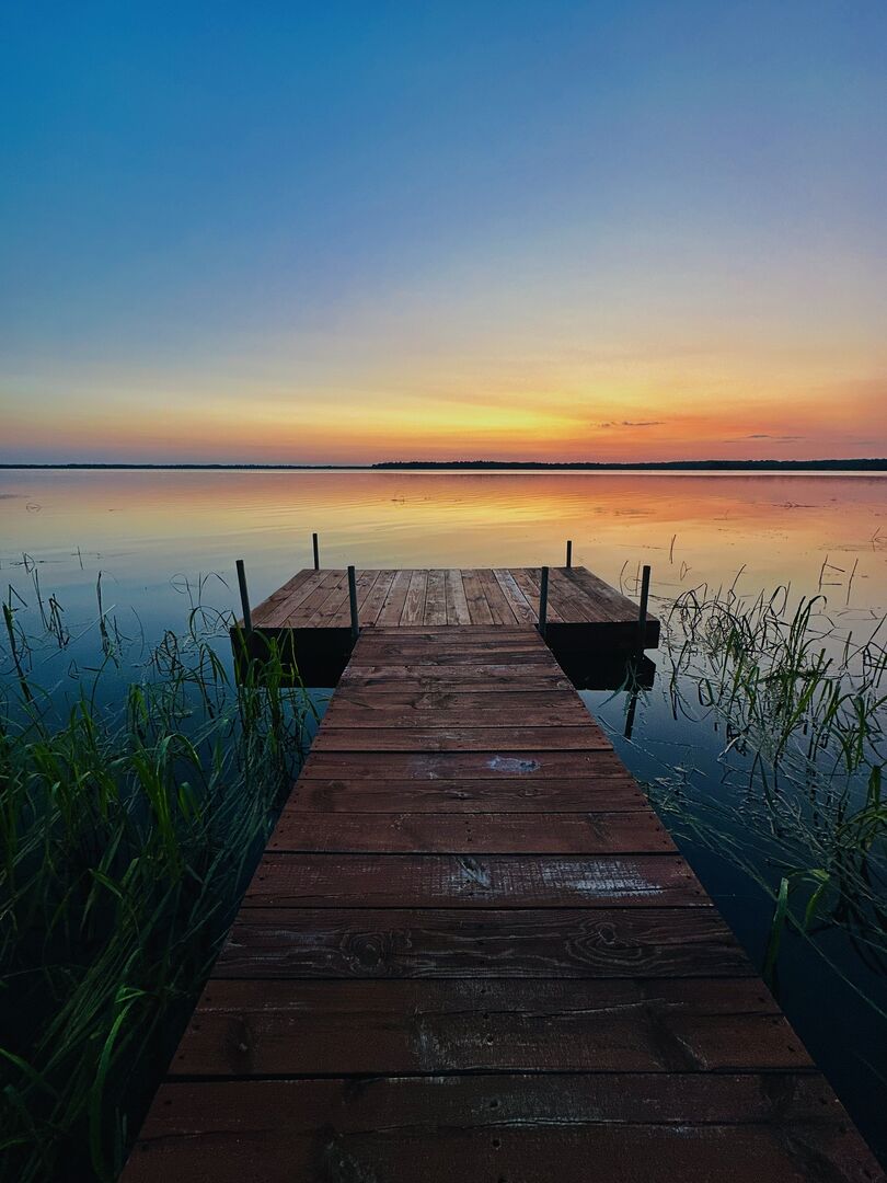 A wooden dock extends over calm waters, with tall grasses peeking out along the edge. The sky is painted in soft hues of orange and blue as the sun sets, creating a serene lakeside scene.