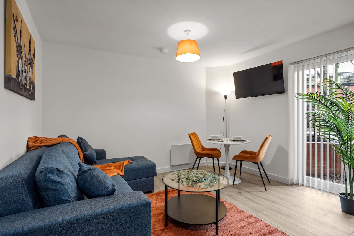 A spacious living room is depicted, featuring a blue sofa complemented by an orange throw. A round coffee table with a decorative centerpiece is arranged on a soft area rug. A dining area with two chairs is visible, and a floor lamp provides warm lighting. Light filters through vertical blinds.