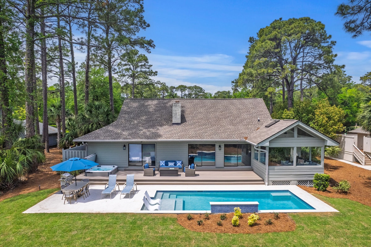 The rear view of the house showcases a spacious deck with outdoor seating and a shaded area under an umbrella. A newly built pool, featuring a tanning ledge, sits adjacent to the deck. Lush greenery and tall trees frame the serene outdoor setting.