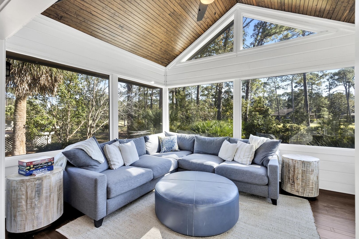A spacious Carolina room features large windows that invite natural light and showcase views of the lush greenery outside. A sectional sofa adorned with decorative pillows and a round ottoman offers comfortable seating, complemented by two side tables. Light wood accents enhance the serene atmosphere.
