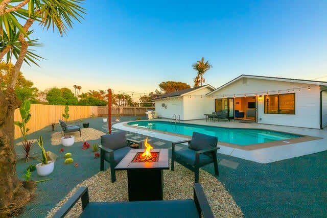 An inviting outdoor space features a glimmering pool surrounded by a stone patio. A cozy fire pit is positioned among four chairs, ideal for relaxing. Lush greenery and gentle lighting accentuate the atmosphere, while a well-maintained home stands in the background.