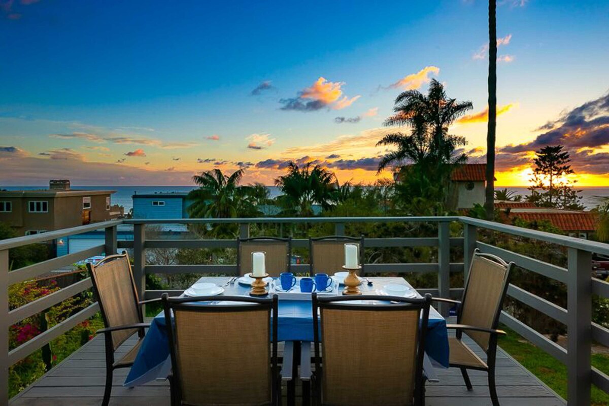 An outdoor dining area is showcased with a table set for a meal, featuring blue placemats and white dinnerware. Surrounding palm trees and colorful foliage provide a natural frame, while the backdrop reveals a vibrant sunset over the ocean.