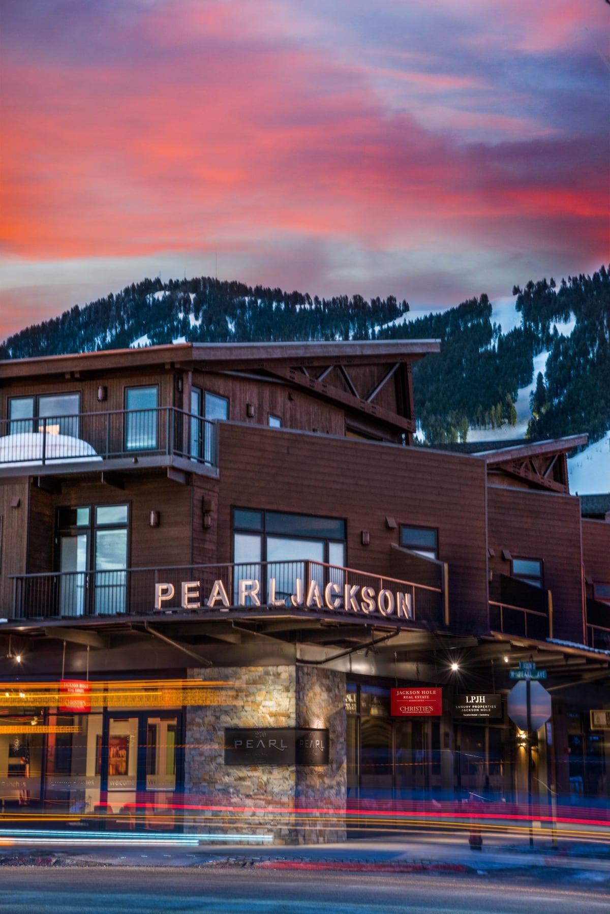 The exterior of the Pearl at Jackson 205 is showcased against a vibrant sunset sky, highlighting its modern architecture. The building features wooden accents and large windows, while its illuminated sign adds a warm touch to the bustling street below.