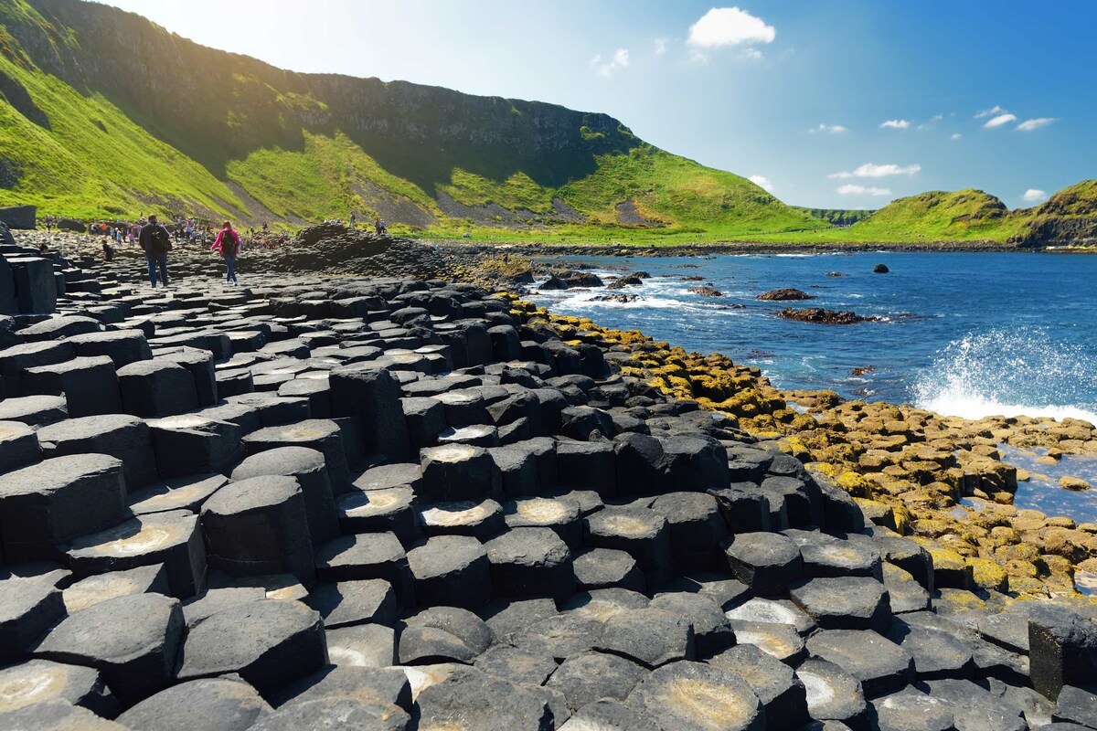 A scenic coastal landscape features distinct hexagonal basalt columns lining the shore. The clear blue water contrasts with the black rock formations, while lush green hills rise in the background under a bright sky. People can be seen walking along the shoreline, enjoying the natural beauty.