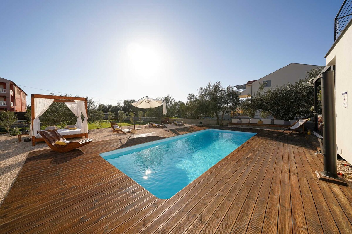 A sunlit pool area features a sparkling blue pool surrounded by a wooden deck. Comfortable lounge chairs are positioned for relaxation, while olive trees add a touch of nature. A shaded seating area with draped fabric offers a cozy retreat beside the pool.