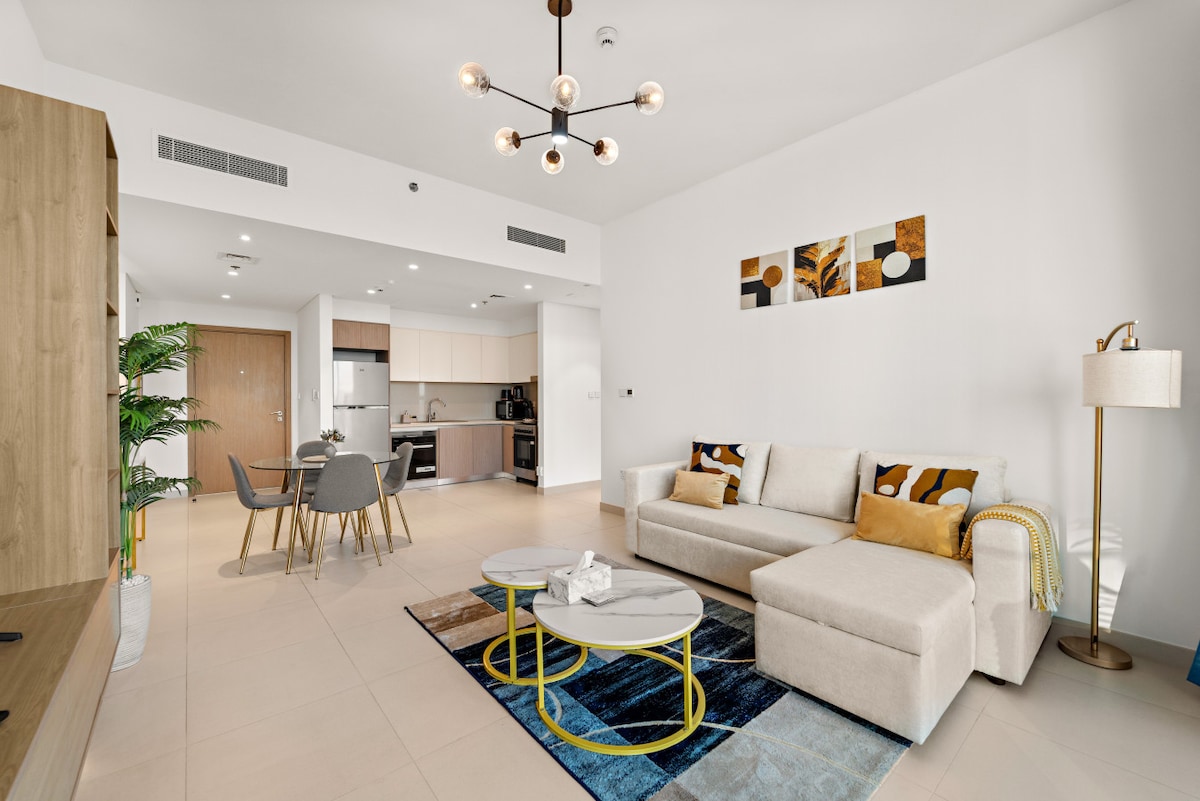 A modern living area showcases a light-toned sofa with accent pillows, accompanied by a coffee table on a blue and yellow rug. A dining area with four chairs is visible in the background, along with a fully equipped kitchen featuring sleek cabinetry and stainless-steel appliances.