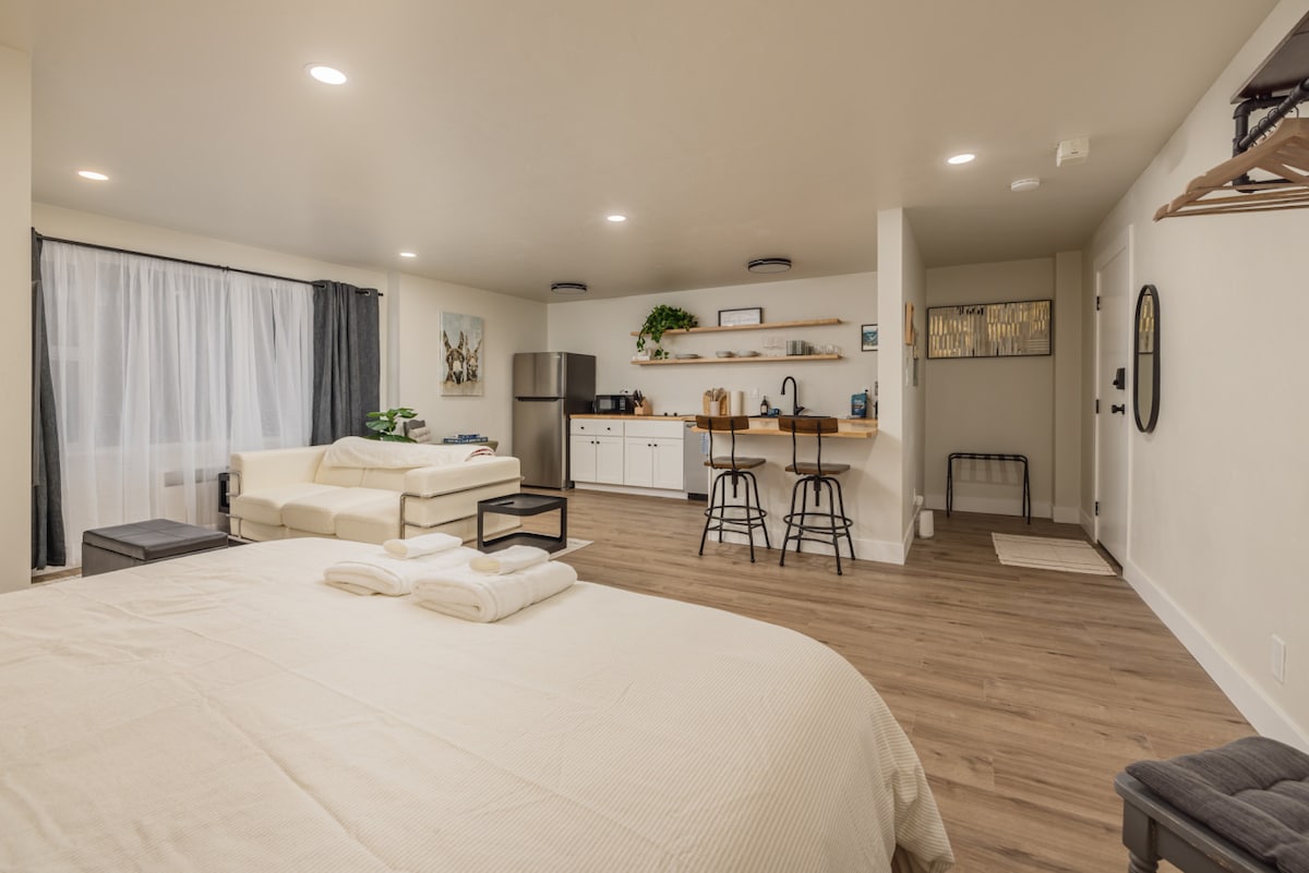 A spacious studio layout is displayed, featuring a king-sized bed with folded towels at the foot. A light-colored sectional sofa is positioned beside a kitchen counter with barstools. Natural light filters through sheer curtains, illuminating modern appliances in the kitchenette.