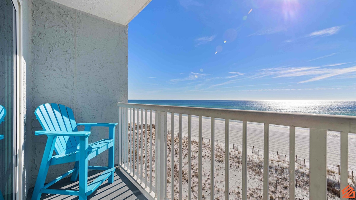 A private balcony offers a view of the shimmering Gulf waters, with sunlight reflecting off the surface. A single brightly colored blue Adirondack chair is positioned at the edge, inviting relaxation. The sandy beach is visible below, enhancing the coastal ambiance.
