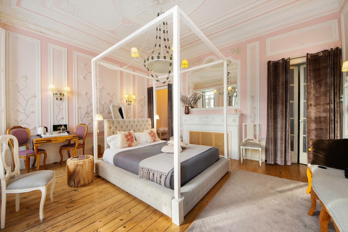 A spacious bedroom is highlighted by a four-poster bed with soft bedding and an accent throw. Elegant wooden flooring complements the pink-walled decor featuring floral motifs. A small seating area is arranged beside a decorative fireplace, with a desk and chairs visible in the background.