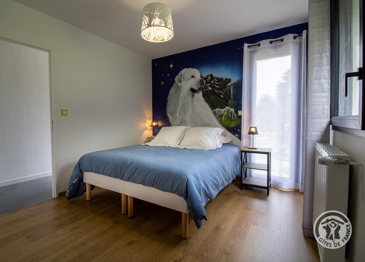 A spacious bedroom features a large bed dressed with soft linens. The wall is adorned with a mural of a white dog under a starry sky. Natural light filters through sheer curtains, illuminating the warm wood flooring and creating a serene atmosphere.