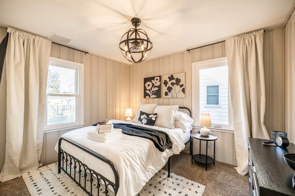 A bright and airy bedroom featuring a comfortable bed with soft linens and decorative pillows. Two windows allow for natural light, complemented by warm lamps on either side. A nightstand and an accent piece with floral artwork add character to the inviting space.