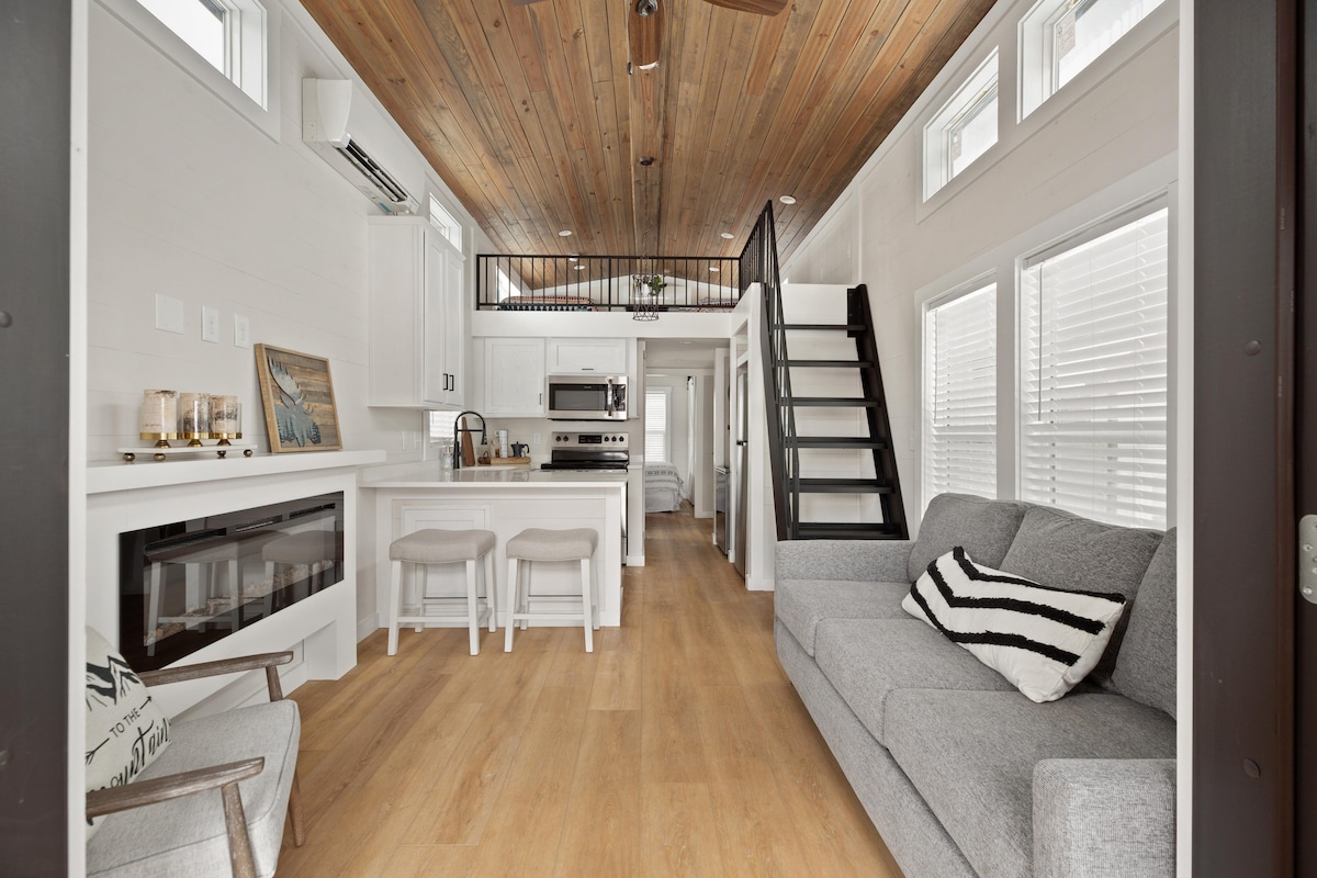 An open living area features light wood flooring and a textured gray sofa with striped pillows. A kitchen island with stools is located adjacent to full-size appliances. Natural light fills the space through several large windows and the warm wood ceiling adds a cozy touch.